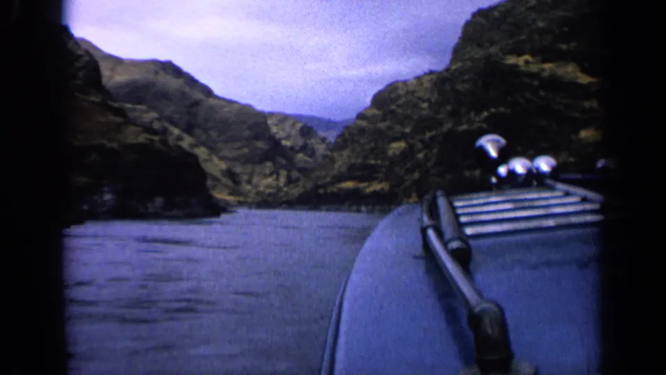 1963 Idaho home-movie frame from the bow of a blue boat traveling between rocky slopes under a clouded sky.