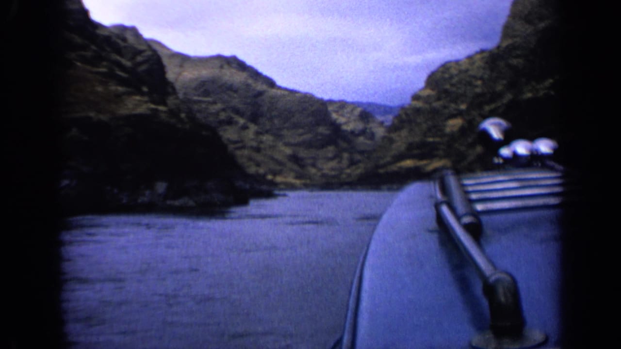 Passenger-view 8mm frame showing the boat foreground, smooth water, and layered rocky slopes along a narrow canyon channel.