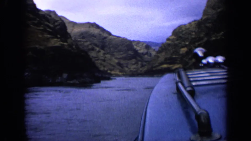Passenger-view 8mm frame showing the boat foreground, smooth water, and layered rocky slopes along a narrow canyon channel.