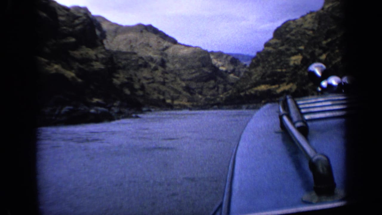 Boat bow crossing calm water between rocky canyon walls under an overcast sky.