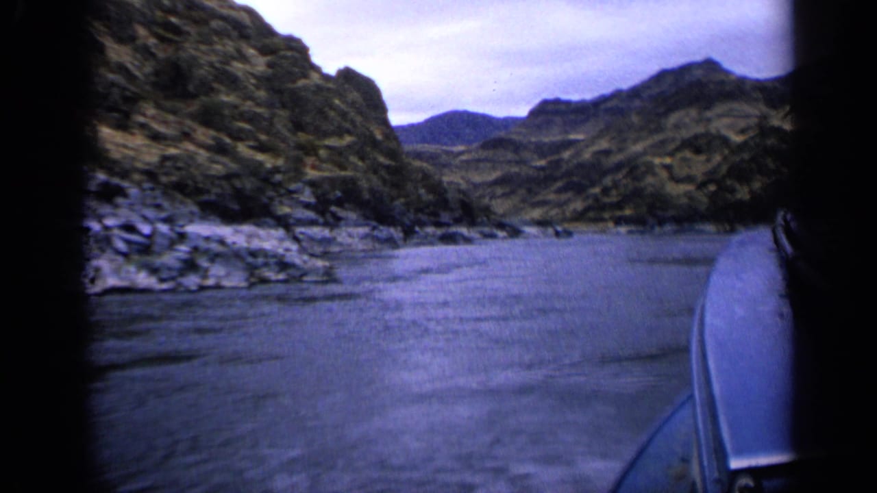Boat approaches calm river water between steep rocky canyon walls under an overcast sky.