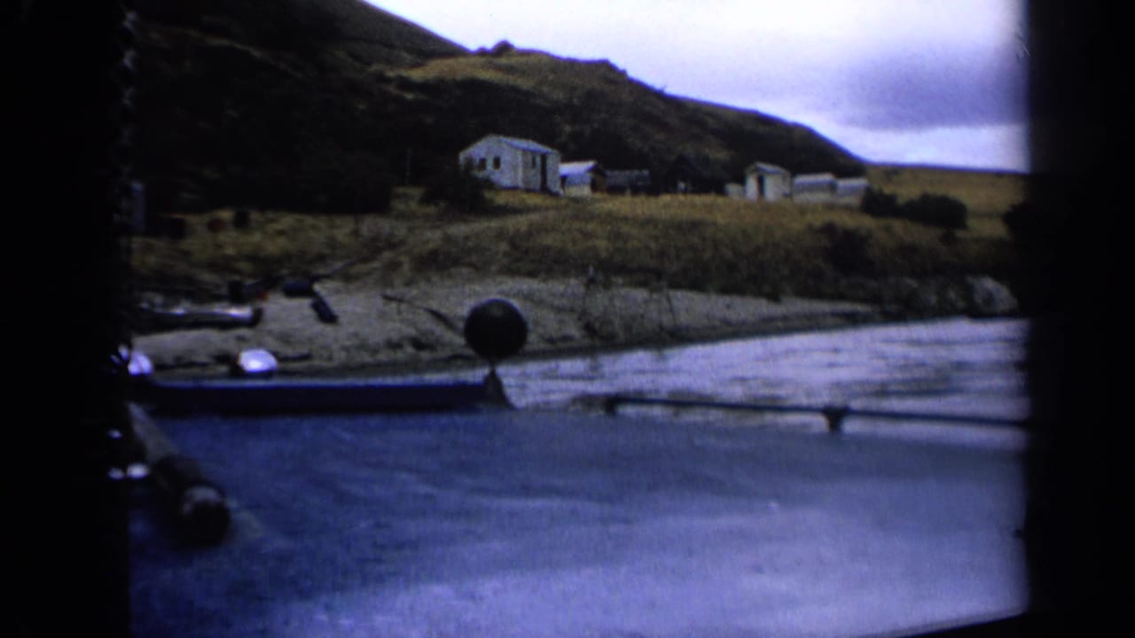 1963 Idaho home-movie frame showing a small riverside settlement, grassy bank and boat hardware in the foreground.