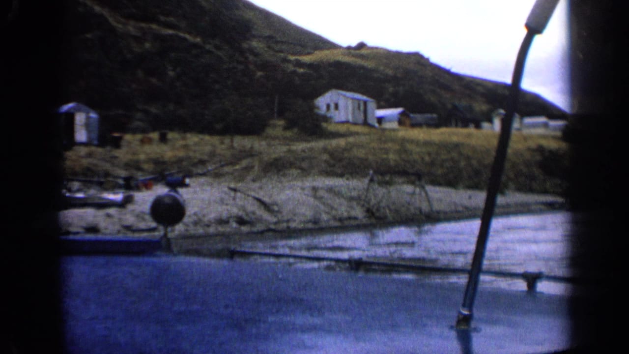 Boat-side view of calm water, grassy shoreline, cabins, and a dark hillside.
