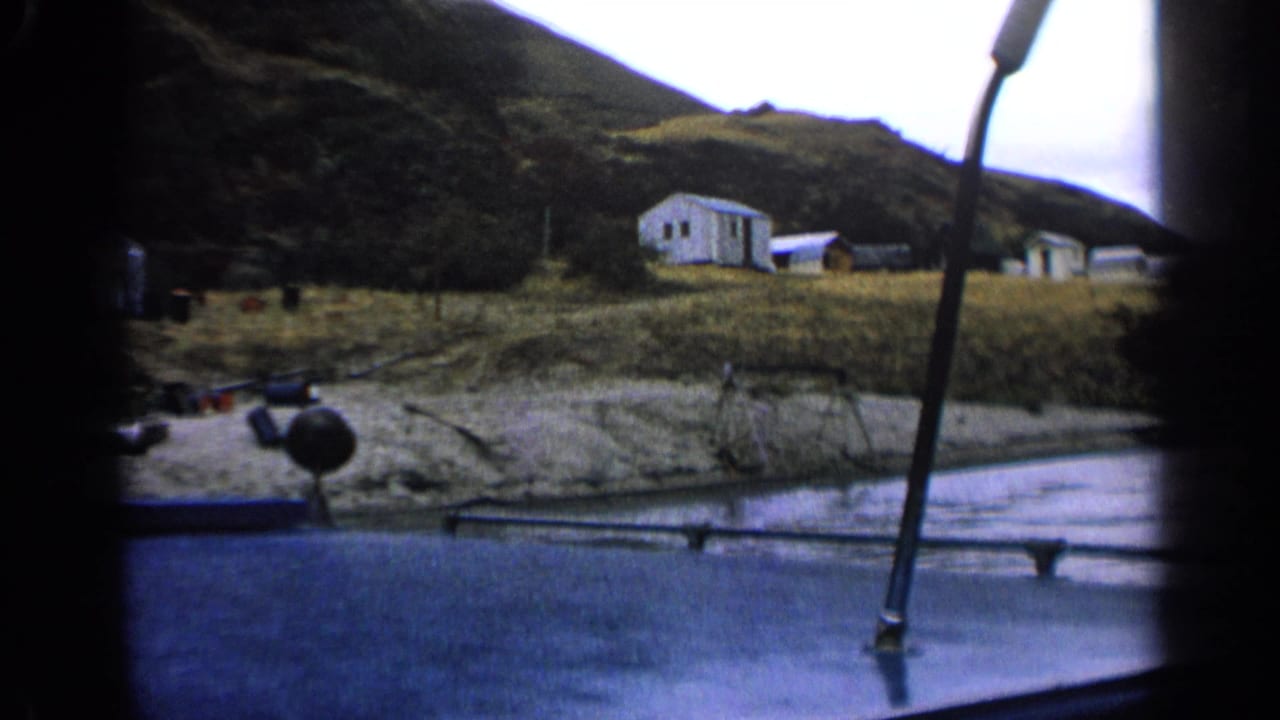 Boat passenger view of Idaho shoreline cabins and grass beneath a rugged hill in 1963 home-movie footage.