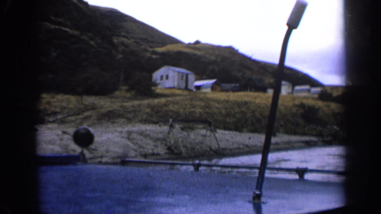 1963 Idaho lakeshore frame showing a white cabin, smaller structures, grass, and a boat rail in front.