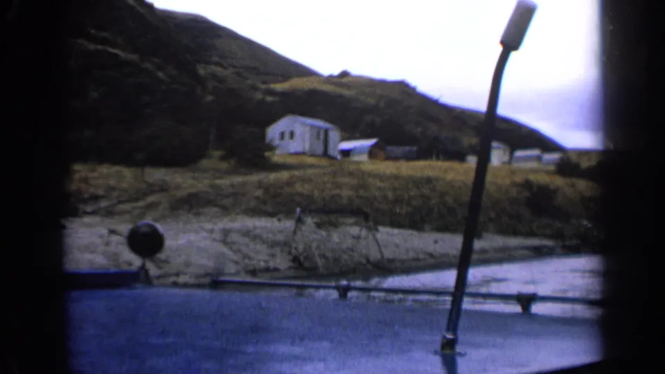 1963 Idaho lakeshore frame showing a white cabin, smaller structures, grass, and a boat rail in front.