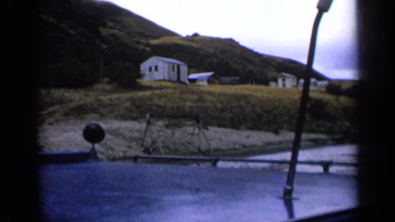 White cabin and small structures on a grassy hillside seen through a vehicle window.