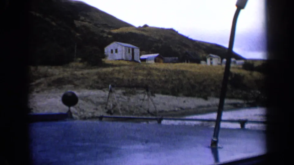 1963 Idaho home-movie view from inside a vehicle toward a white cabin, smaller outbuildings, grass, and dark hillside under a pale sky.