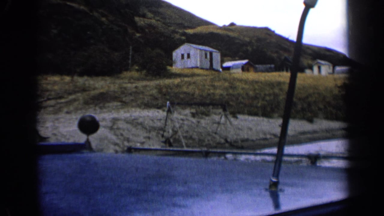 A vehicle-window view in 1963 Idaho frames a small white building, nearby structures, grass, and a dark hillside in soft 8mm color.
