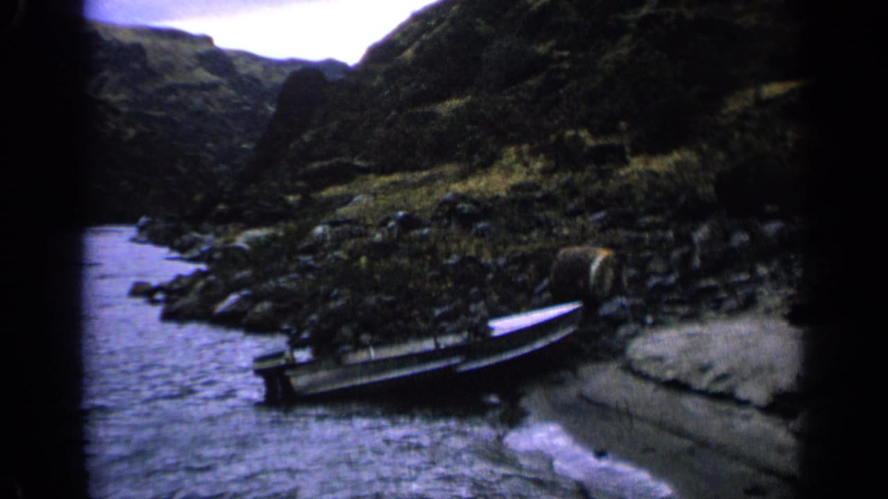 A 1963 Idaho riverbank scene shows a small boat pulled onto rocks beside moving water, with rugged hills rising behind it.