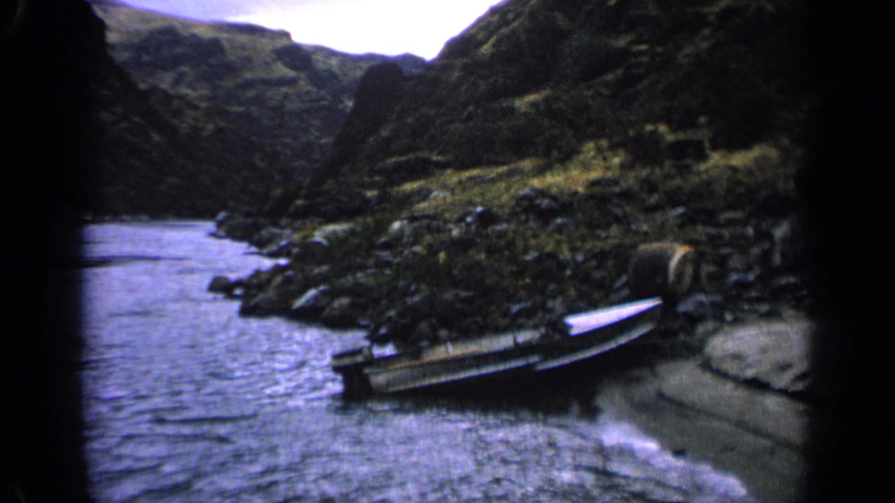 The later frame holds on the 1963 Idaho riverbank, with a small open boat resting on rocks near the water and steep hills beyond.