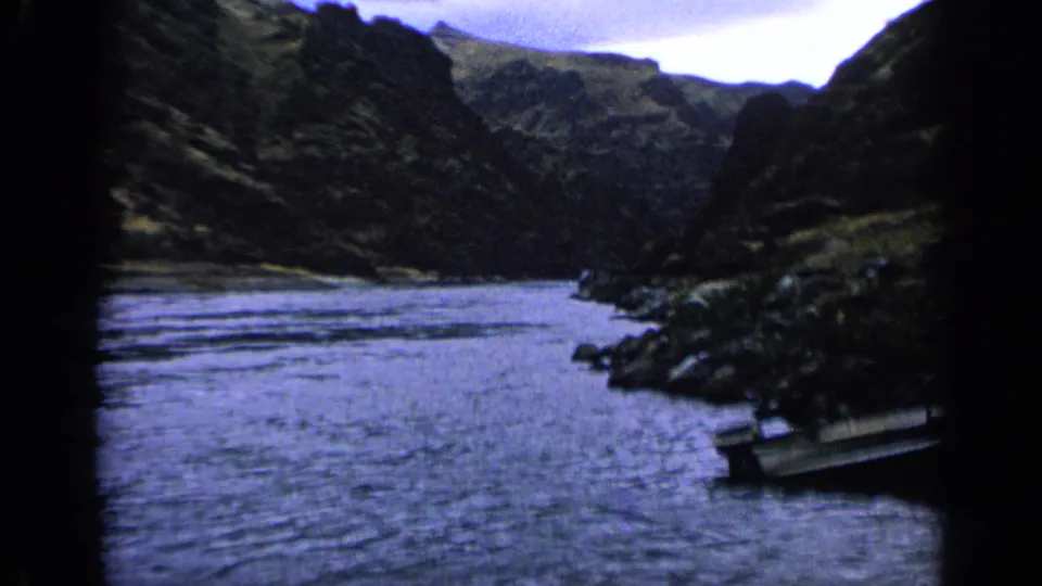 1963 Idaho 8mm view across a broad stream toward steep hills, with a partly visible boat at the lower right edge.