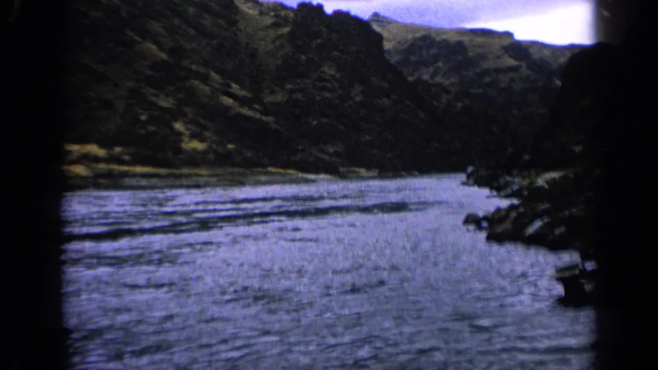 1963 Idaho home-movie frame of a broad stream between steep valley walls, with the boat nearly out of view at the lower right.