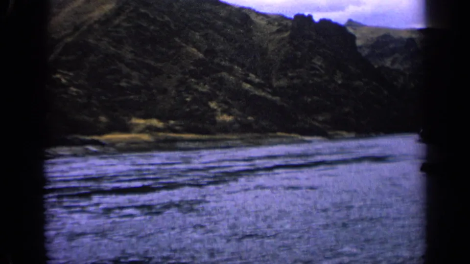 Idaho home-movie frame from 1963 with rippled river water, rugged hillsides, and a muted overcast sky.