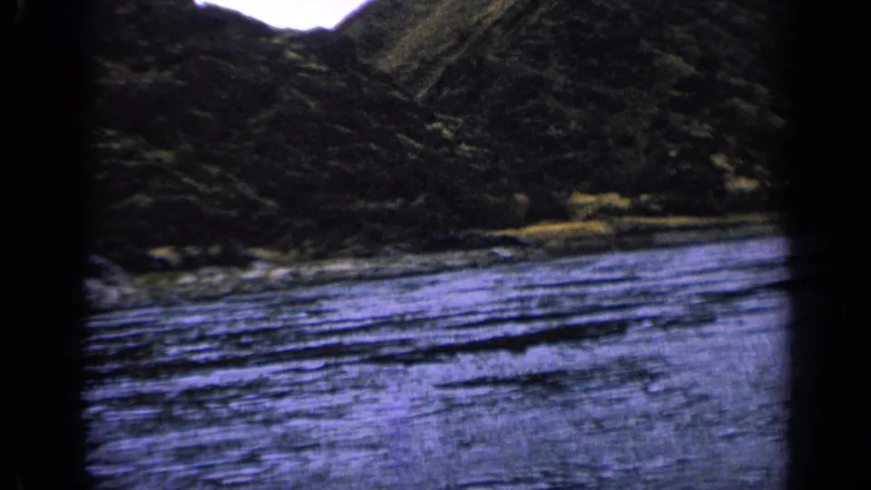 Final 1963 Idaho frame with textured river ripples, sloped rocky terrain, and visible 8mm grain and color shift.