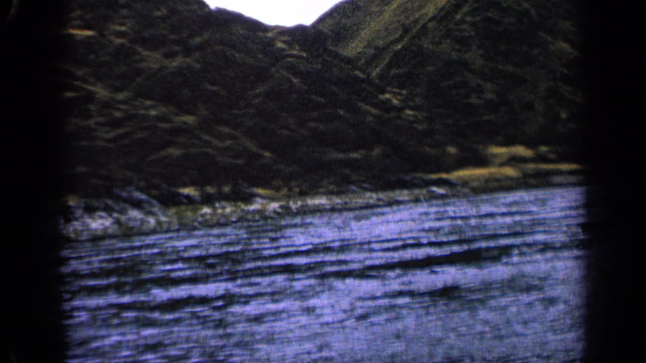 1963 Idaho 8mm frame showing rippled river water, rocky bank, dark hillside, and black film border from a moving viewpoint.
