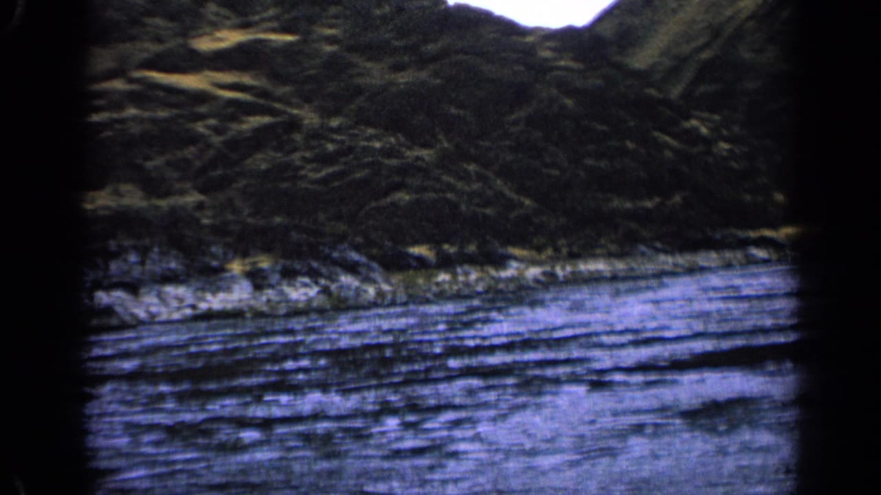 River and rocky bank seen beneath brush-covered hills.