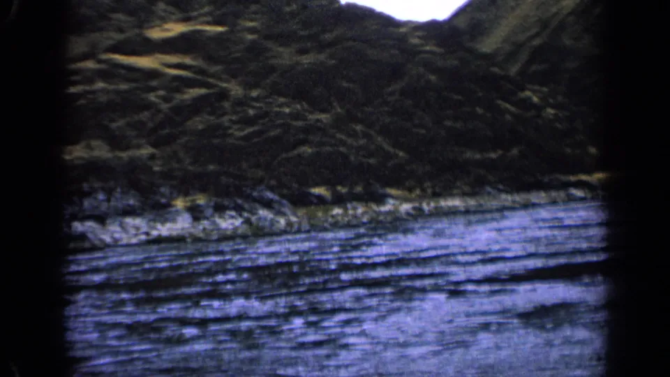 1963 Idaho home-movie frame with blue-purple river water, rocky shoreline, brush, hills, and visible 8mm gate edges.