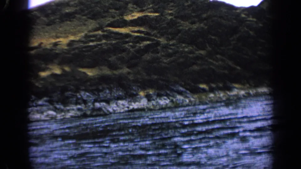 Moving 8mm view in Idaho, 1963, with a rippled river, rocky shoreline, dark hillside, sparse vegetation, and soft focus.