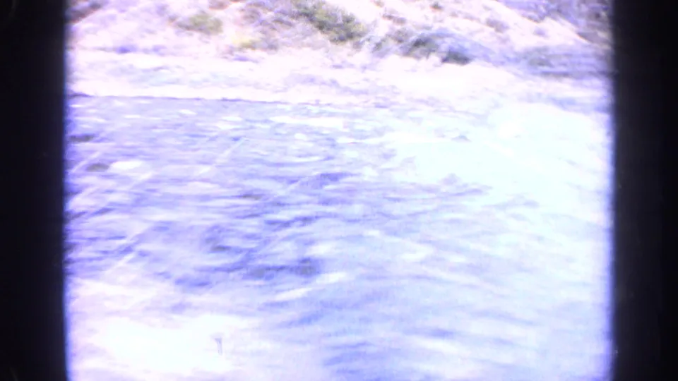 1963 Idaho 8mm frame showing bright overexposed river water, distant rocky bank, pale shoreline, and dark film edge.