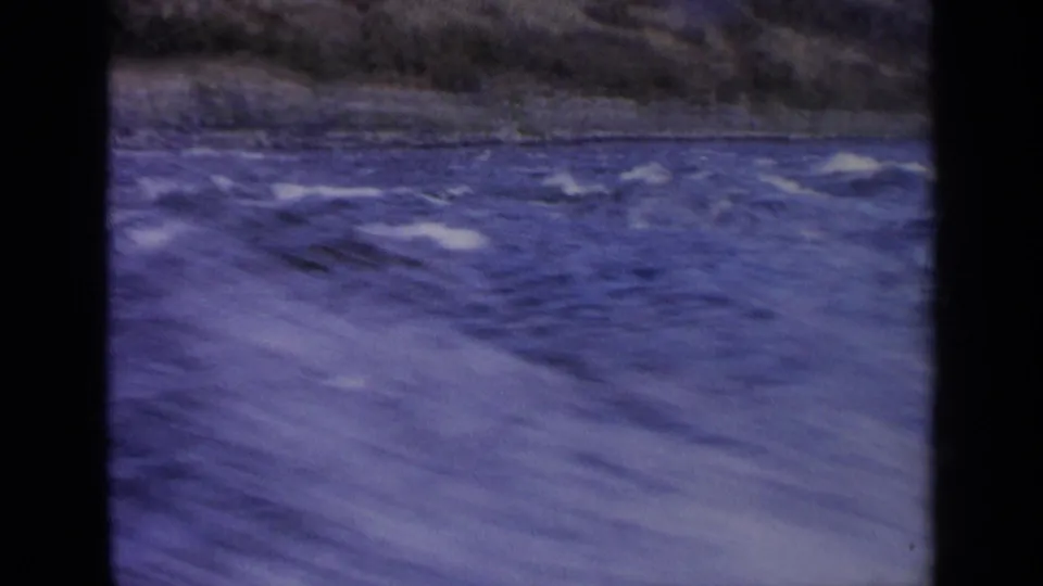 1963 Idaho 8mm frame with fast-moving river water, pale foam, soft motion blur, and a dark rocky riverbank.