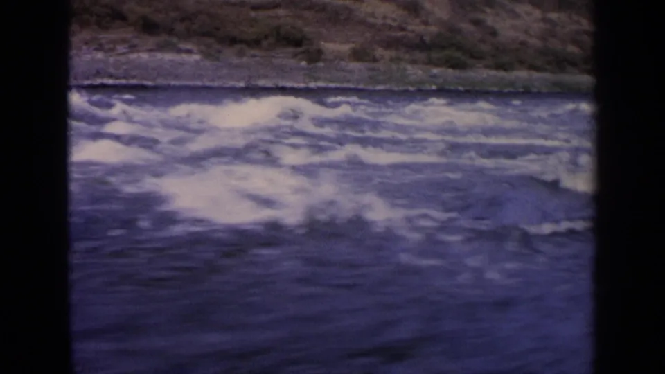 1963 Idaho 8mm frame with whitecaps on blue river water and a rugged, sparsely vegetated bank beyond.