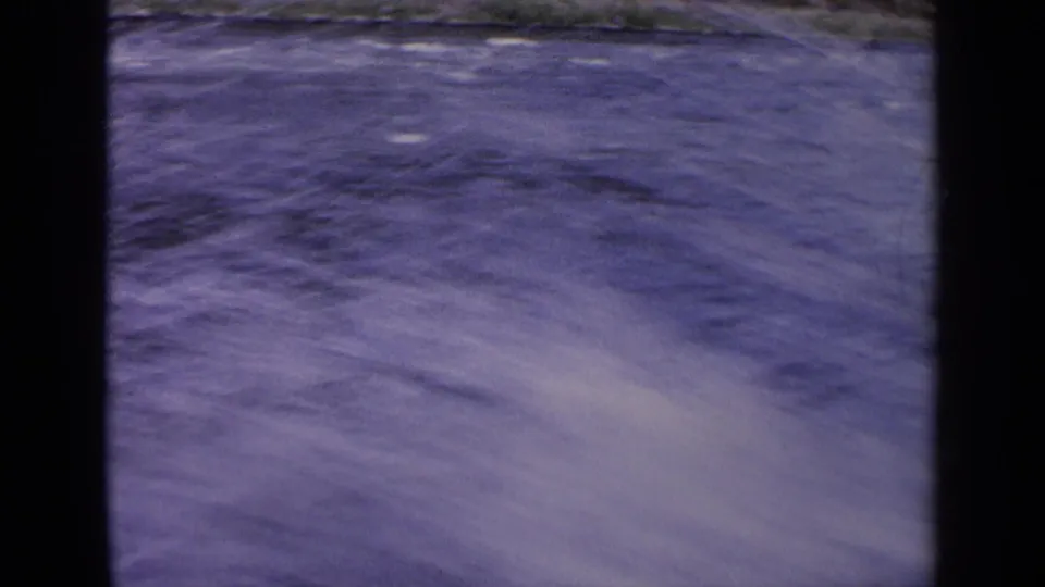 1963 Idaho 8mm frame with motion-blurred blue river water, pale foam, and a narrow strip of rocky shoreline near the top.