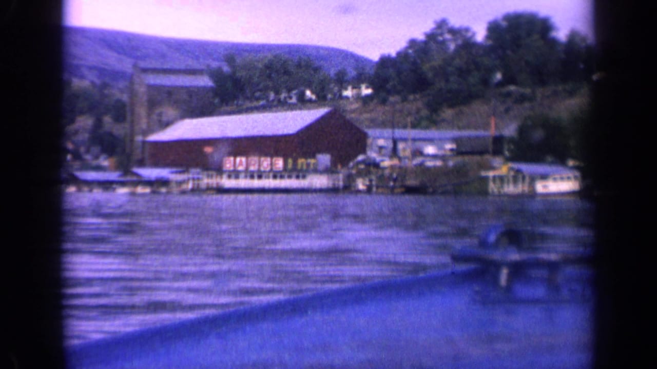 Red shoreline building, docks, boats, trees, and hills across a lake.