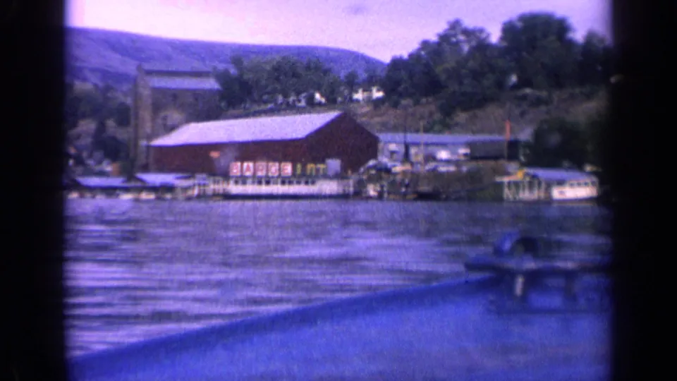 1963 Idaho lake view from a boat, with a red barn-like shoreline building, docks, small boats, trees, and low hills.