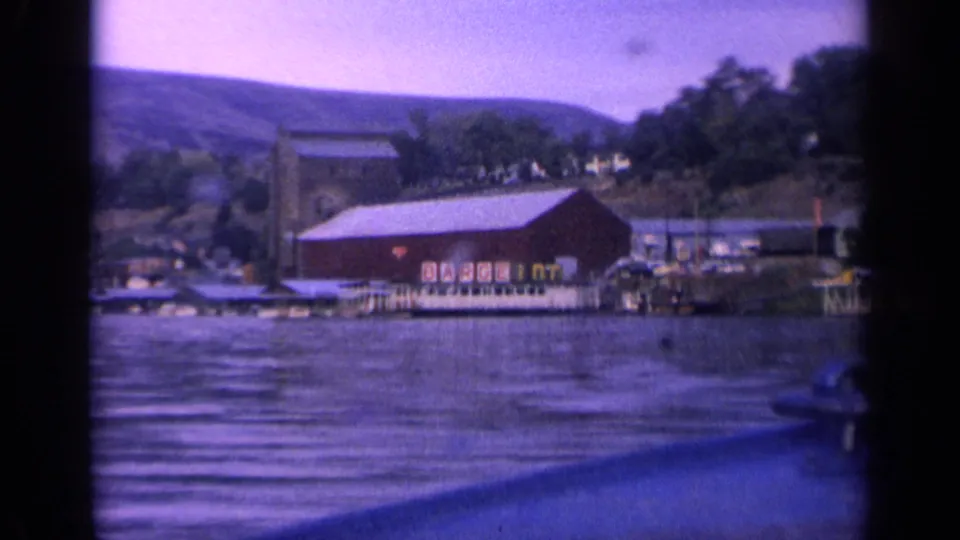 Boat-level 8mm view of a red lakeside building and dock area in Idaho, with trees and hillside houses in the background.