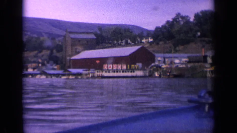 1963 Idaho home-movie frame showing a red shoreline building, dock, wooded bank, distant hill, and the edge of the filming boat.