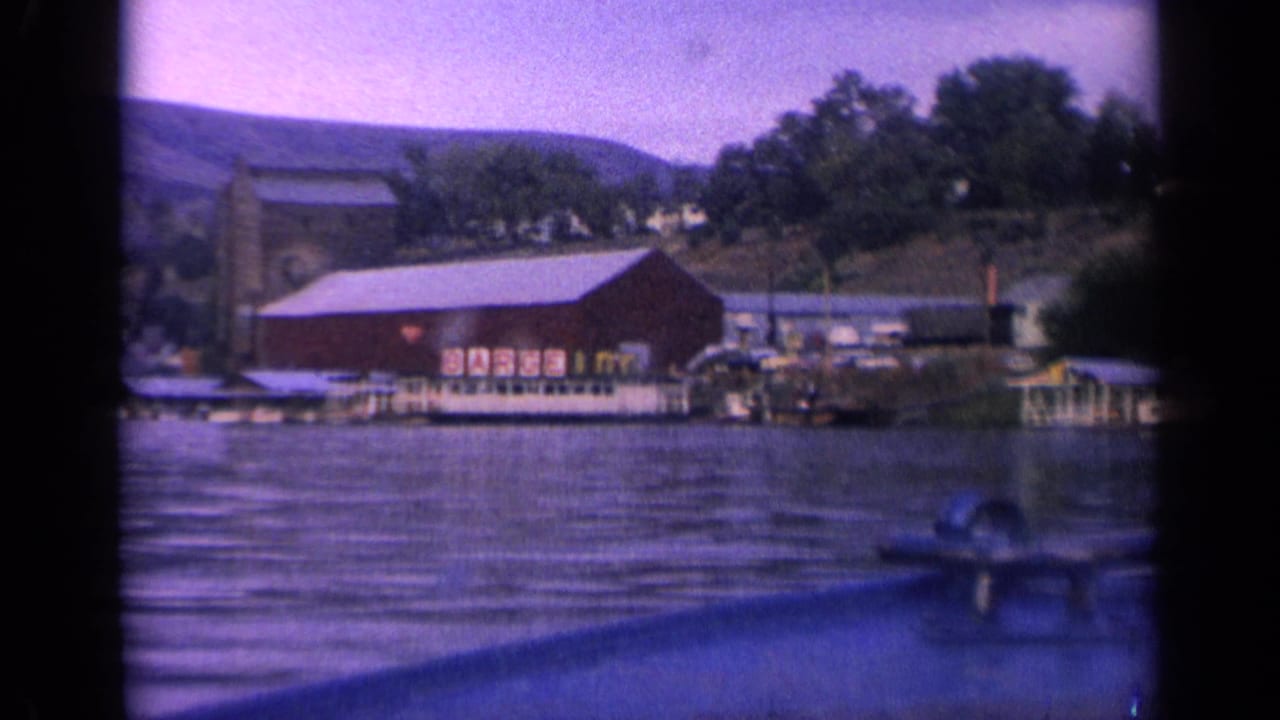 1963 Idaho 8mm frame showing a red waterfront building with BARGE INN lettering, docks, hills, and lake ripples viewed from a moving boat.