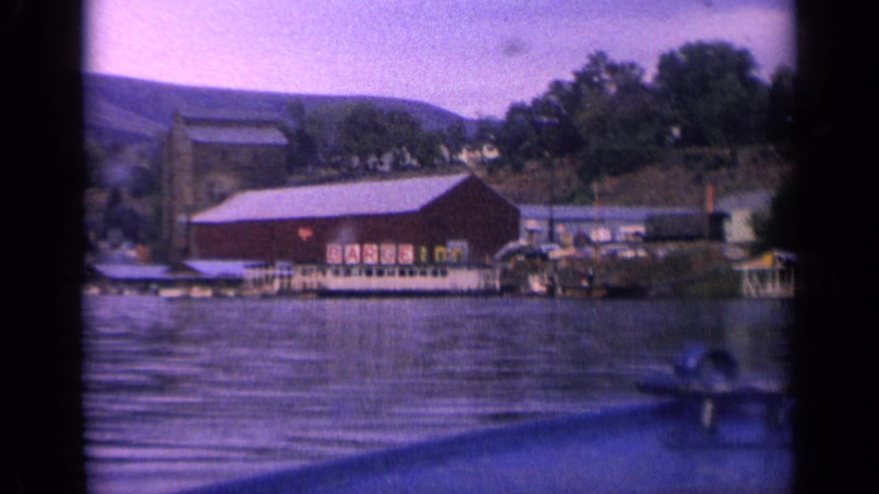 1963 Idaho 8mm view of the BARGE INN shoreline building, docks, trees, and hillside across blue-purple lake water.