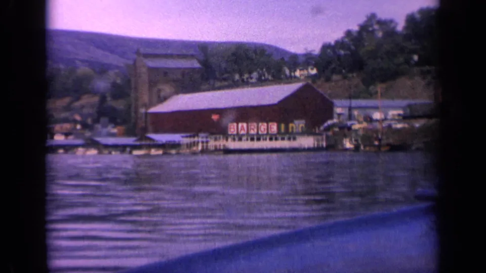 Soft 8mm frame from 1963 Idaho showing the red BARGE INN building beside docks, with rippled lake water and wooded hillside behind.