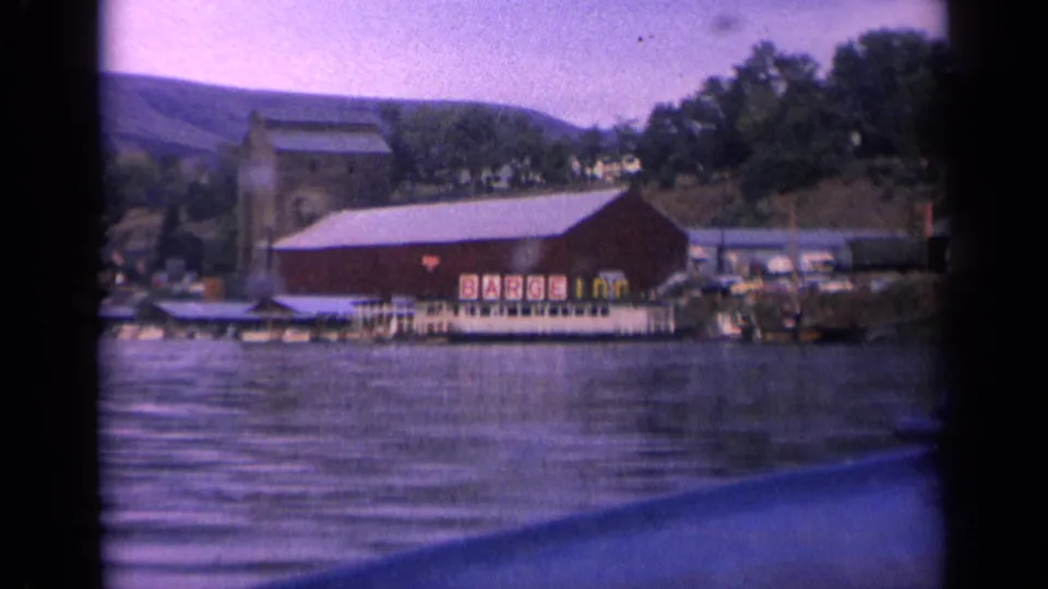Final 1963 Idaho 8mm frame with the BARGE INN building, dock area, trees, hillside, and dark boat edge crossing the foreground.