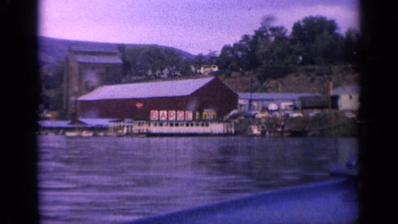 1963 Idaho 8mm frame showing a red lakeside inn-like building, dock, lake water, trees, and hillside through a projected film frame.