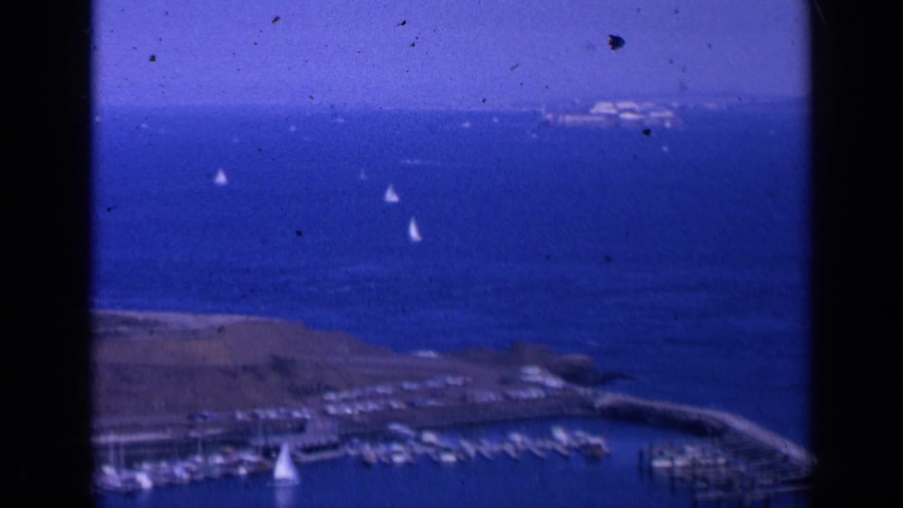 Blue bay water with several white sailboats, a marina below, distant land, and film gate edges.