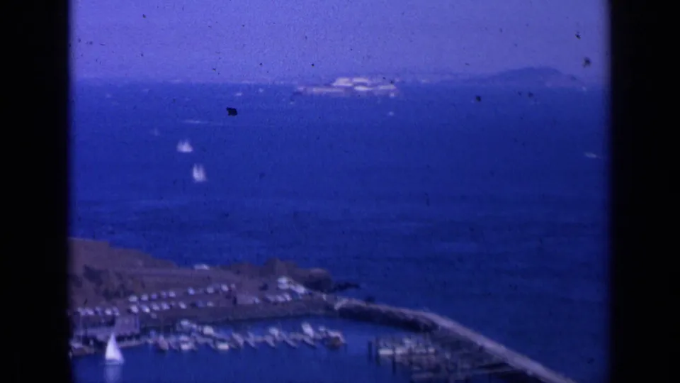 1963 home-movie frame looking over a San Francisco Bay marina, with docked boats, open blue water, small sailboats, and distant shoreline.