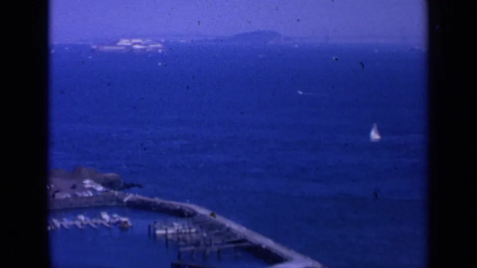 Aerial 8mm frame of San Francisco Bay with a lone sailboat on blue water, marina docks at the bottom, and distant land on the horizon.