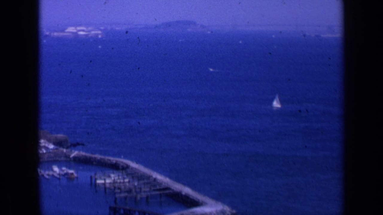Blue bay water with a small white sailboat, pier, distant shoreline, and dark film borders.