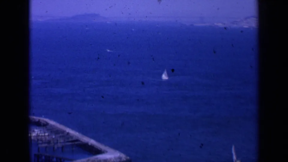 Elevated 1963 San Francisco bay view showing a small sailboat, blue water, partial pier, and distant shoreline.