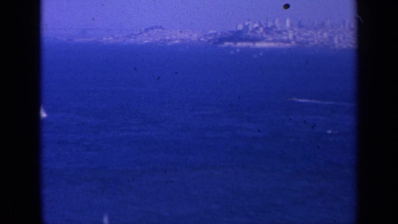 1963 home-movie frame showing San Francisco across deep blue bay water, with a hazy skyline and visible 8mm frame edges.