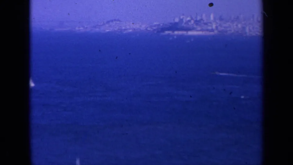 1963 home-movie frame showing San Francisco across deep blue bay water, with a hazy skyline and visible 8mm frame edges.