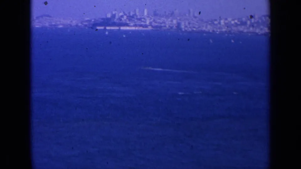 Distant San Francisco skyline over blue bay water, with a pale wake crossing the mid-distance in a grainy 8mm frame.