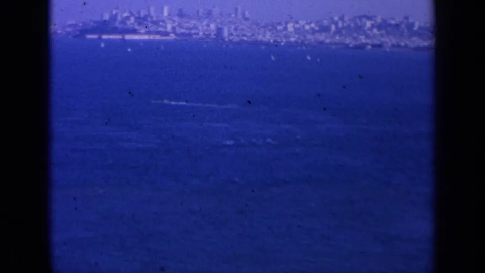 1963 San Francisco bay view with the city on the horizon, small boats, and white wakes visible through blue-tinted 8mm film.