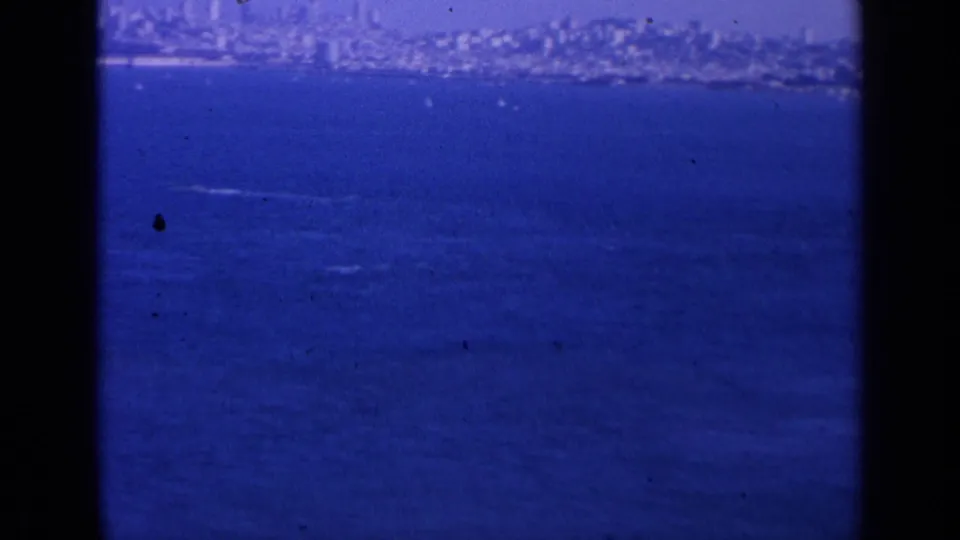 Blue-tinted 8mm view of San Francisco across the bay, with a small wake on the water and the skyline softened by haze.