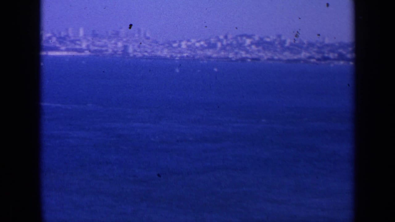 Distant 1963 San Francisco cityscape seen across blue water, with hazy buildings, shoreline, and visible 8mm film borders.