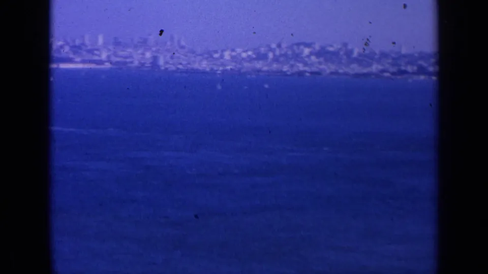 Distant 1963 San Francisco cityscape seen across blue water, with hazy buildings, shoreline, and visible 8mm film borders.