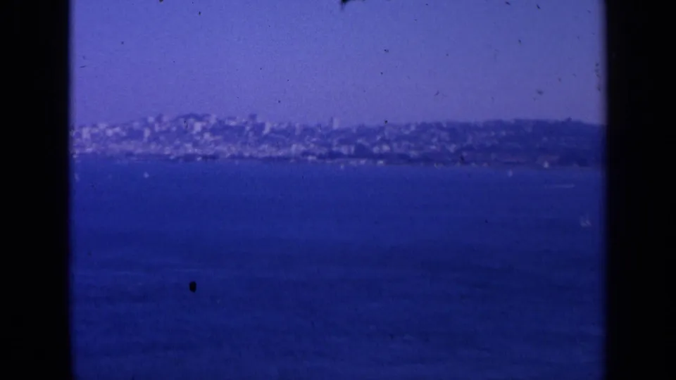 A soft 1963 home-movie view looks across blue water toward a distant San Francisco shoreline and hillside cityscape.