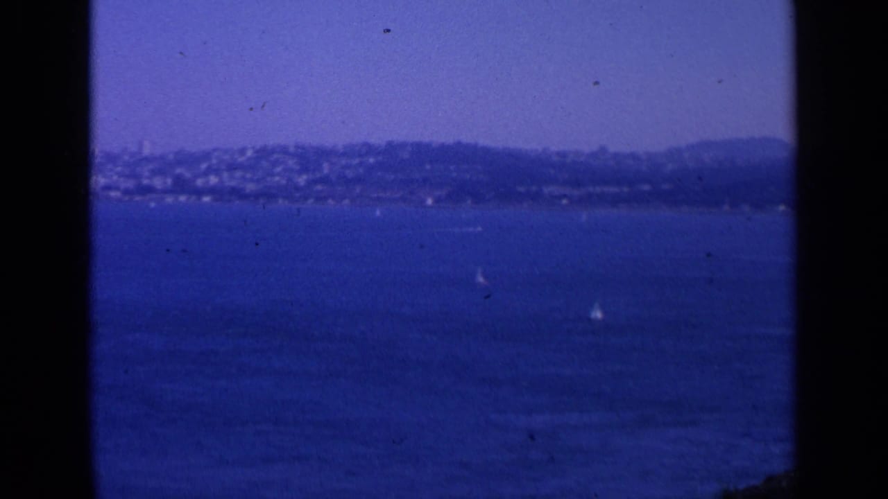Blue bay water with distant hills and small sailboats under a clear sky.